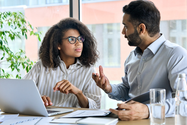 Indian CEO converses with African American colleague, both using a laptop. Multicultural professionals gather in a boardroom, discussing a business plan.