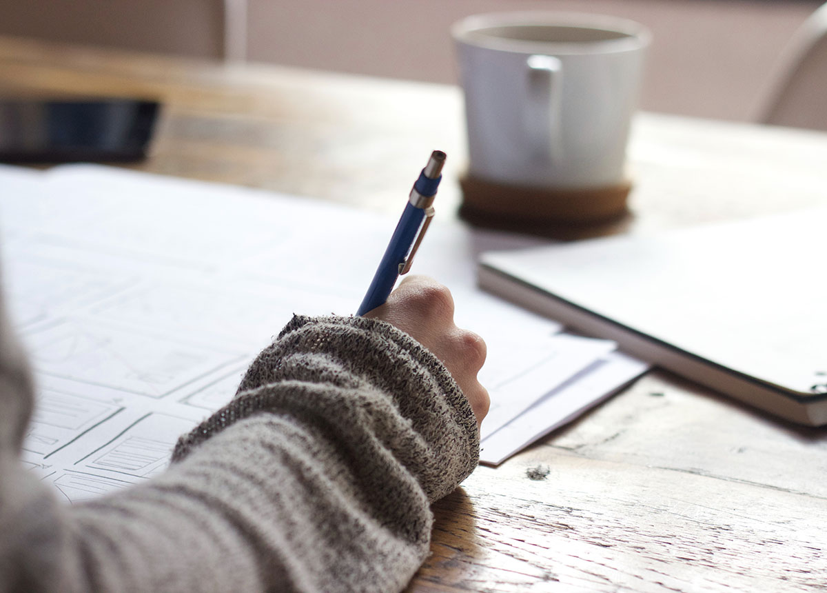 person's hands writing in journal with a coffee in the background