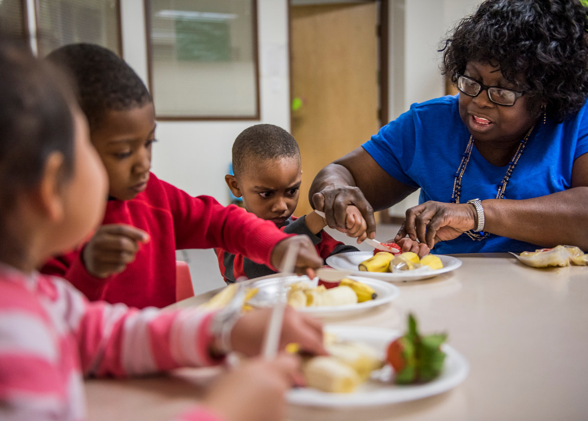 woman helping child cut fruit with plastic knife