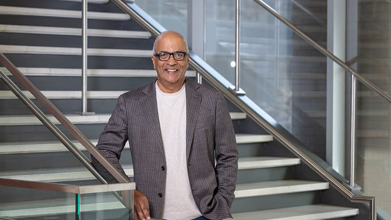 Madhavan Parthasarathy in a gray suit jacket and white t-shirt standing at the bottom of a staircase wearing dark, square-rimmed glasses and smiling with his teeth.