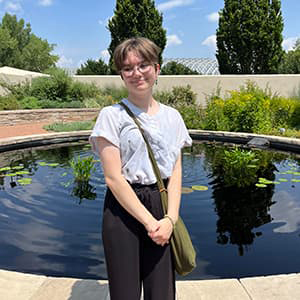 Kiera Tomlinson stands near a pond in black pants and a white blouse looking off to the side with short hair and glasses.