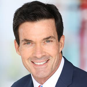 Scott Webb wearing a navy suit with a tie and white shirt, short dark hair, and smiling with his teeth.