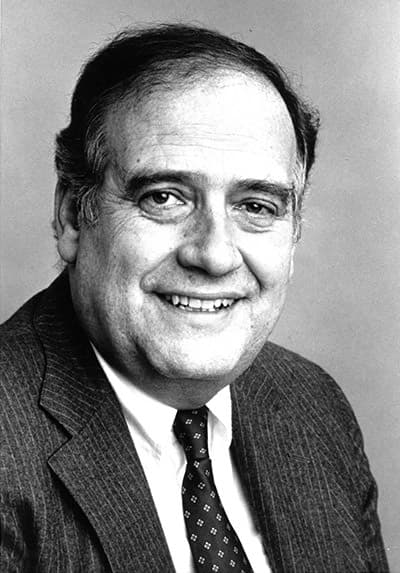 Black and white Dwayne Nuzum wearing a suit and tie with sort hair and smiling with his teeth.