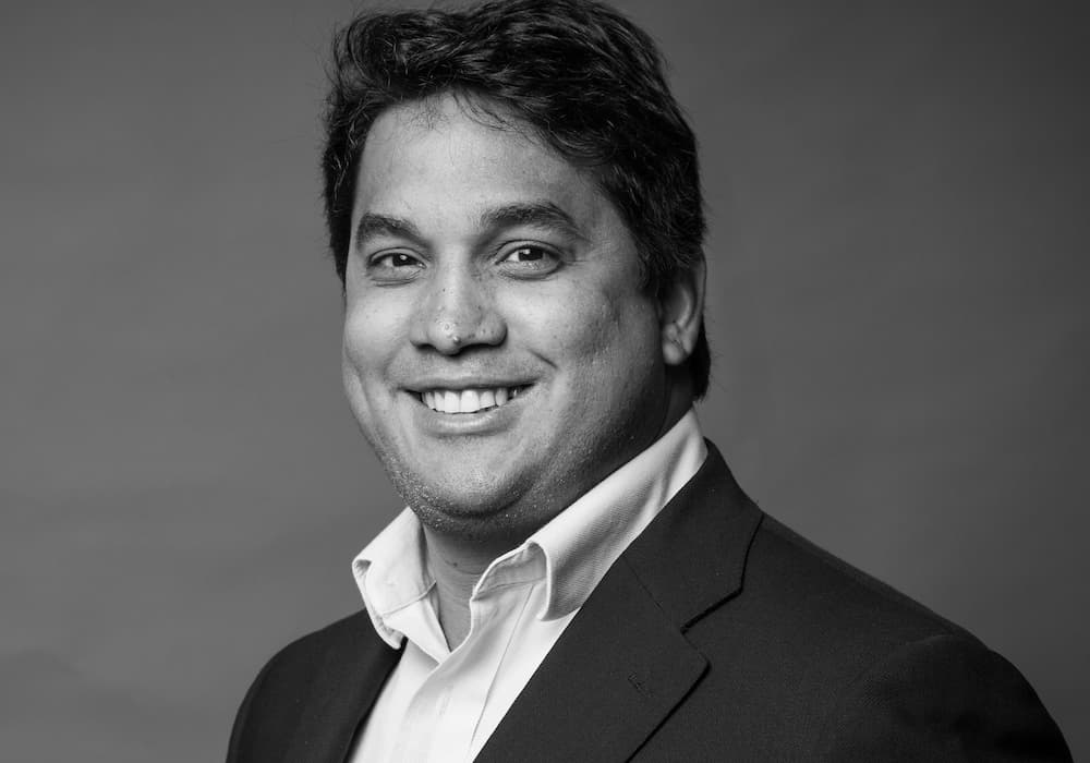 Black and white photo of Hugo Azevedo's face and shoulders wearing a white button up and dark jacket, smiling with his teeth.