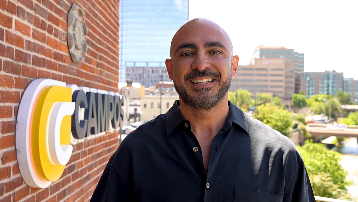 Marco Campos wearing a black polo and smiling with his teeth standing outside against a brick wall with Campos branding.