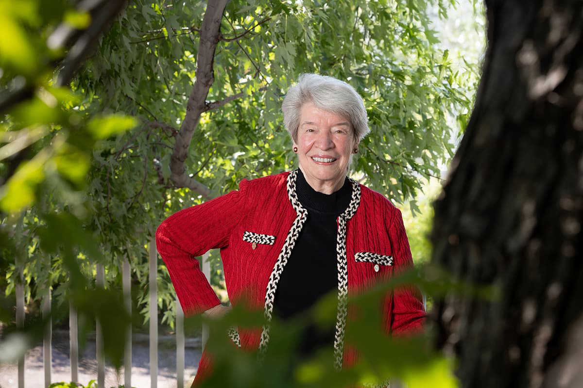 Mary Guy stands outside in a red cardigan with short white hair smiling with her teeth.