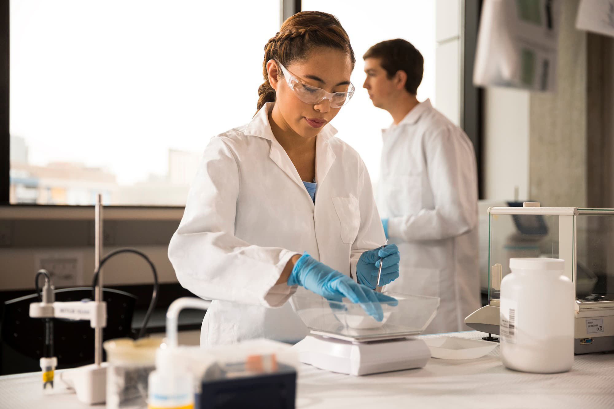 Student wearing a white lab coat, blue gloves, and safety glasses working in a lab. Student wearing a white lab coat, blue gloves, and safety glasses working in a lab.