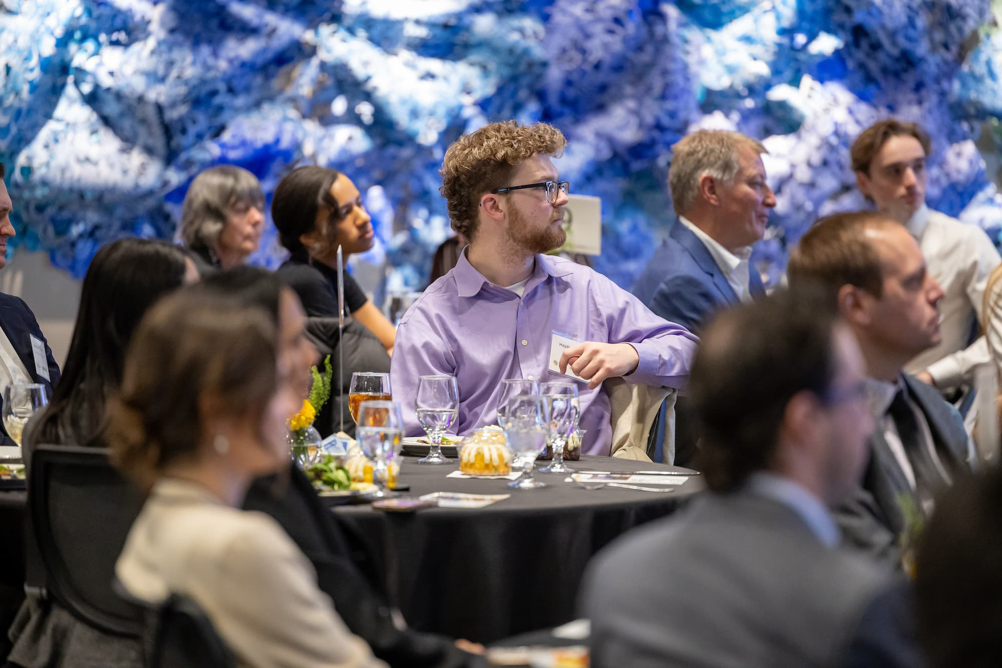 A crowd of students seated at round tables dressed in business attire looking toward the same direction.