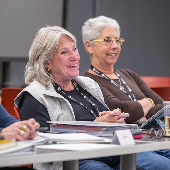 Two mature women seated at a classroom table smiling.