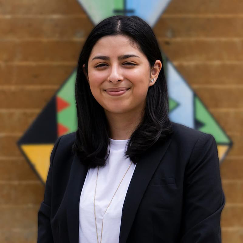 CHloe Frazee pictured from her ribs up with a mouth-closed smith. She's wearing a white shirt and dark suit jacket and stands in front of a brick wall.