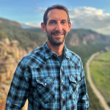 Dan Ben-Horin stands outside wearing a black and blue plaid shirt, short brown hair and beard, smiling with his teeth.