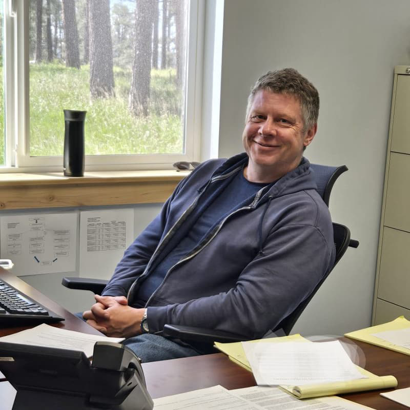 Guy Patterson casually seated at a desk covered in papers, leaning back and smiling. He wears a blue t-shirt and zip-up hoodie.