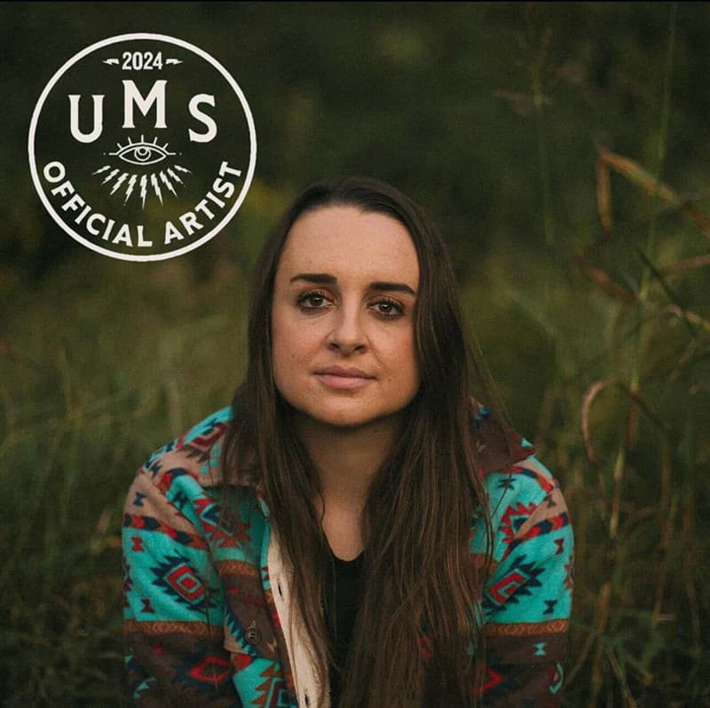 Olivia Rudeen in a colorful blouse and long, dark hair, seated in a field of long green grass.