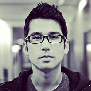 Simon Moya-Smith black and white portrait, wearing dark, square-rimmed glasses, and short dark hair.