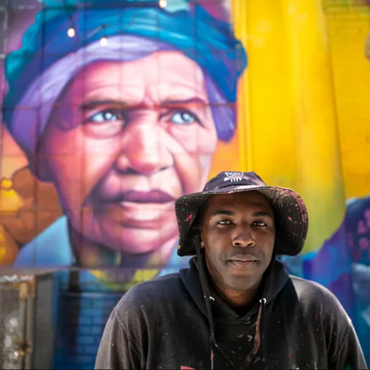 Detour sits in front of one of his colorful murals dressed in black and covered in paint smiling.