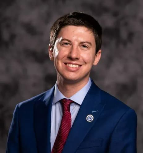 Image of William Lindstedt smiling with his teeth in a blue suit and red tie, gray background.