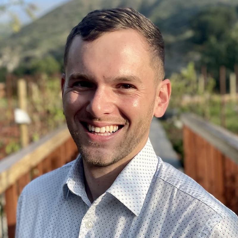 Zachary Lees from the shoulders up in a white pattered button-up shirt, smiling with teeth, outdoors.