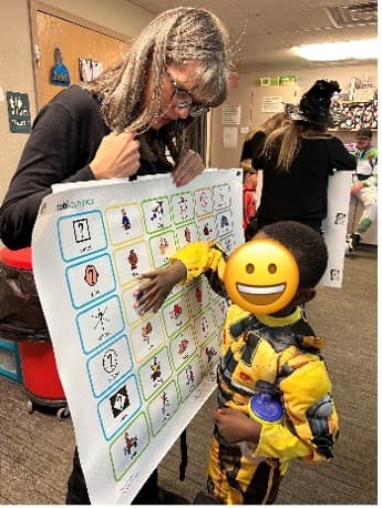 teacher with child pointing to picture symbolsAlt text: classroom of children with teacher pointing to communication board